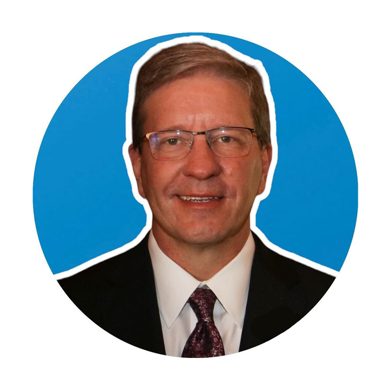 A man wearing glasses, a dark suit, white shirt, and patterned tie smiles in a portrait with a blue background.